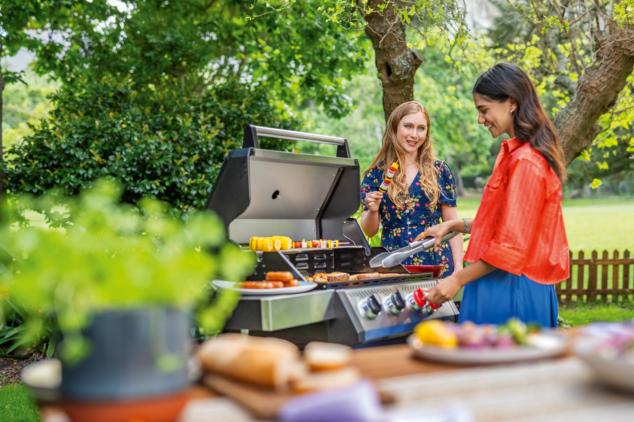Två kvinnor grillar korv, majskolvar och grönsaksspett utomhus.