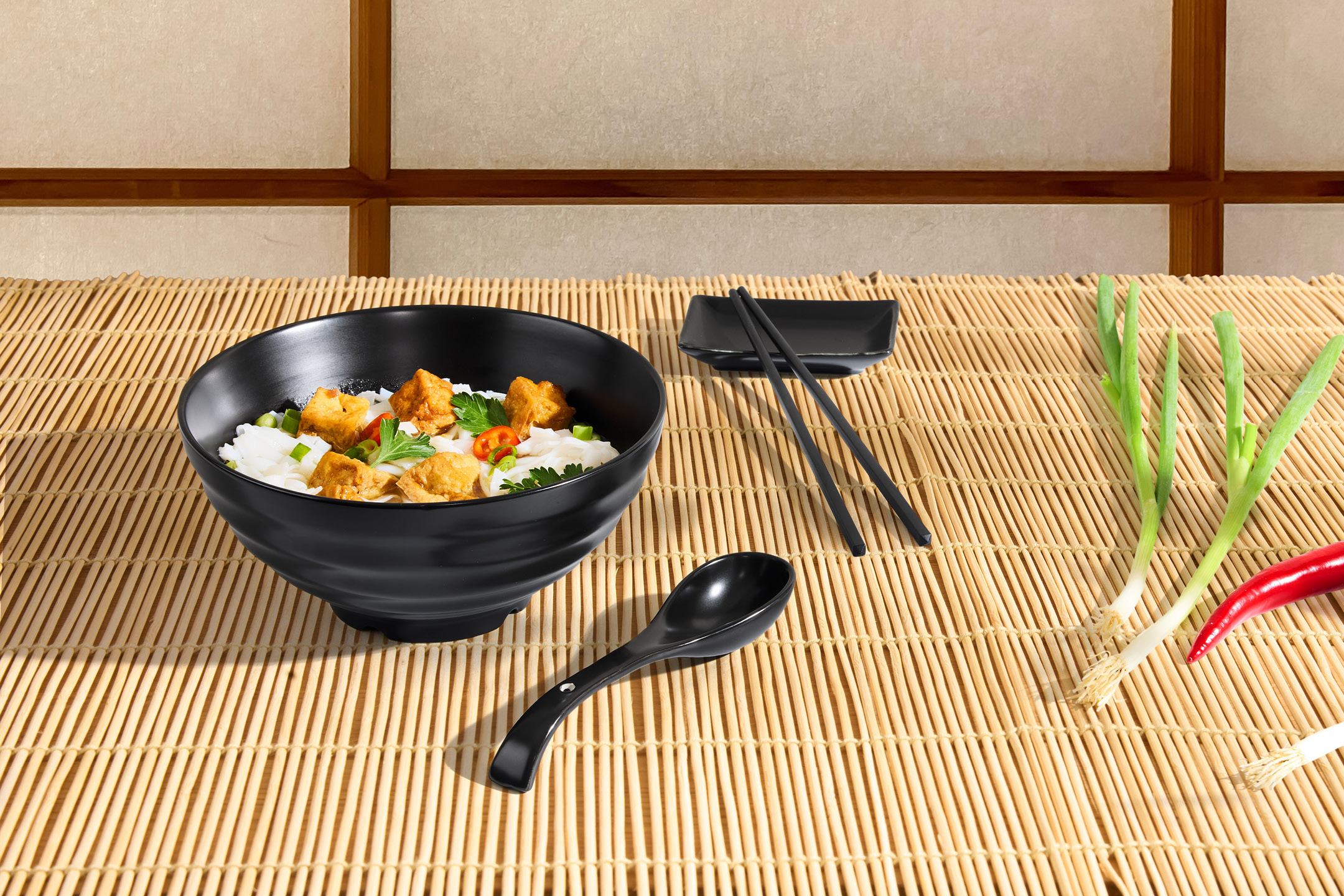 En skål med risnudlar och tofu, ätpinnar, sked, salladslök och chili på en bambumatta.