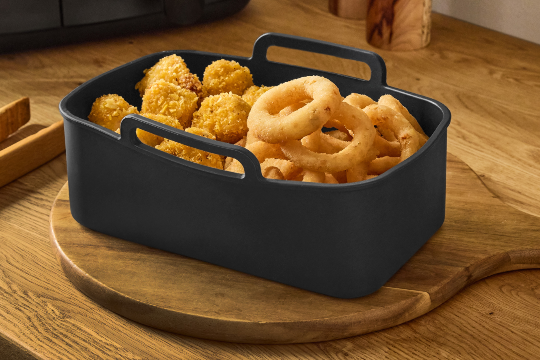 En svart silikonform fylld med gyllene lökringar och panerade kycklingbitar på en träskärbräda.