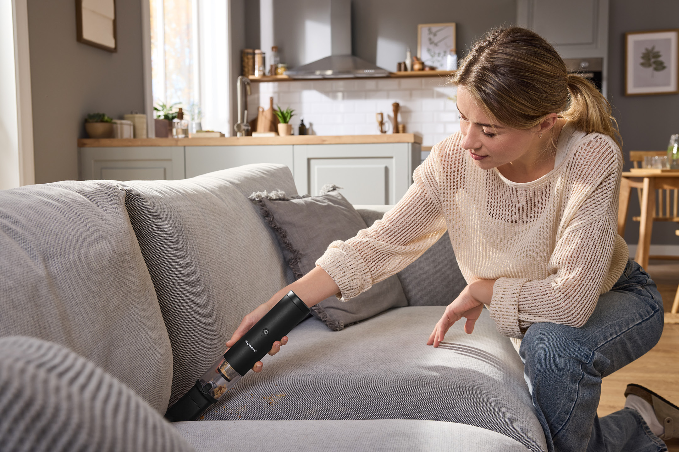 Kvinna dammsuger en soffa med en handdammsugare i ett ljust vardagsrum.