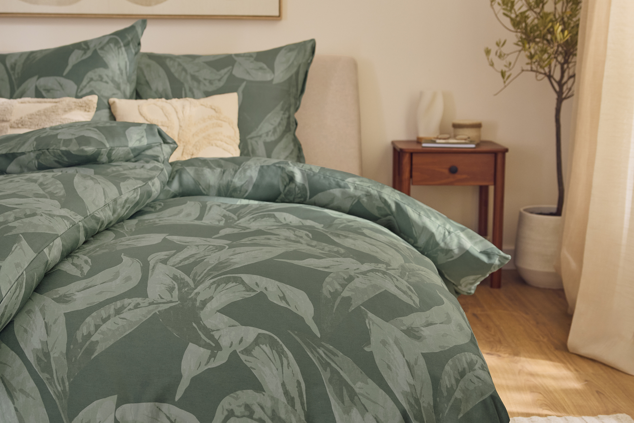 Sängkläder med grönt bladmönster i ett sovrum med nattduksbord och växt.