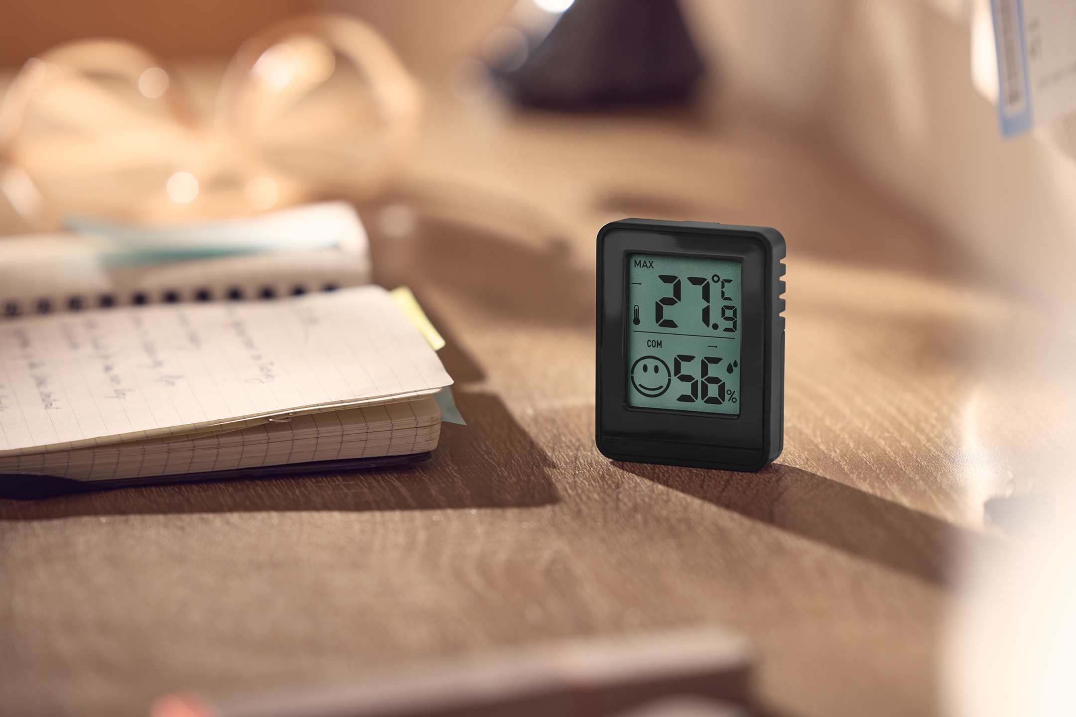Digital termometer och hygrometer på ett skrivbord med en anteckningsbok.