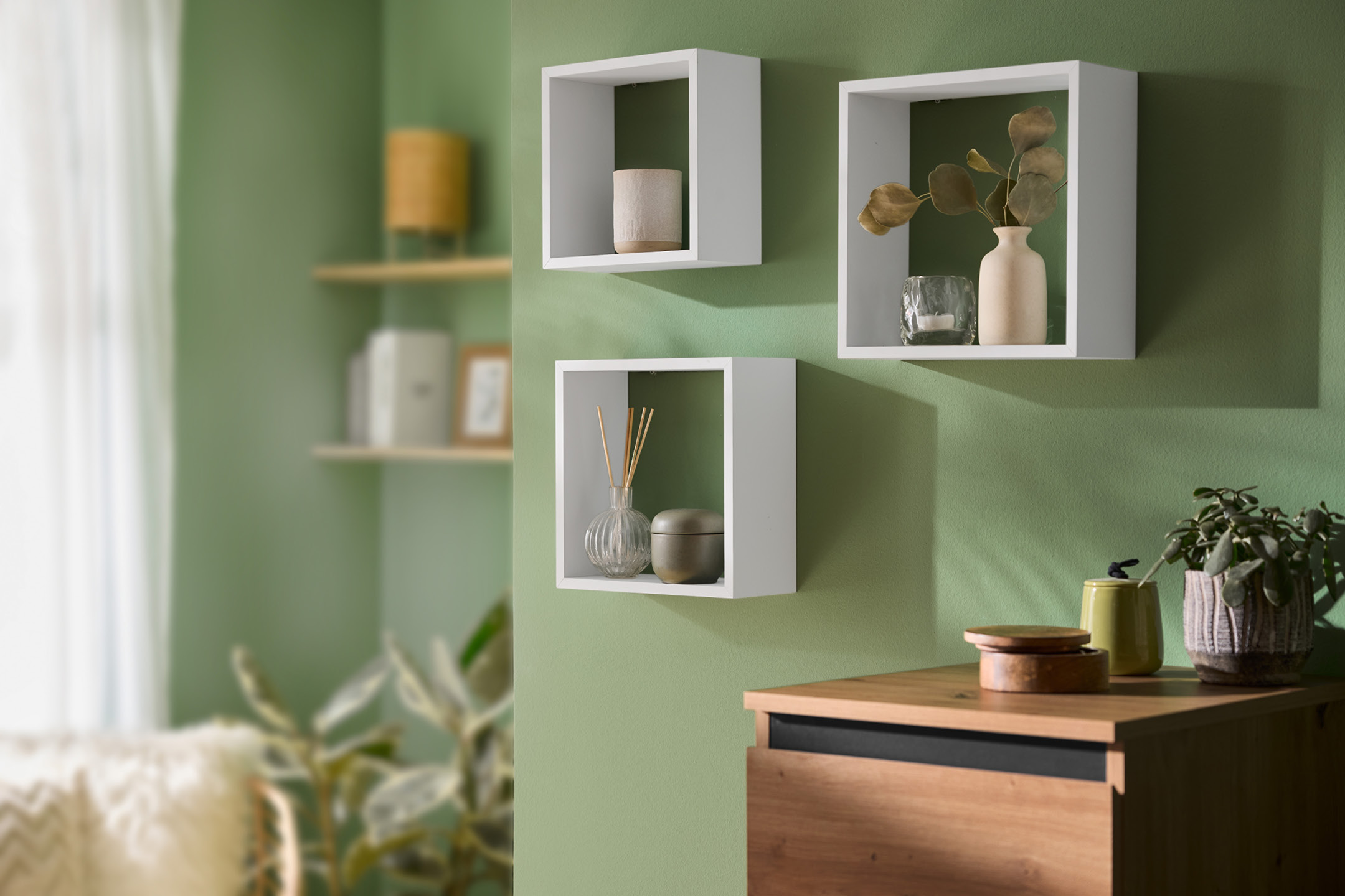 Väggmonterade vita kubhyllor med dekorativa föremål och en träbyrå med en växt.