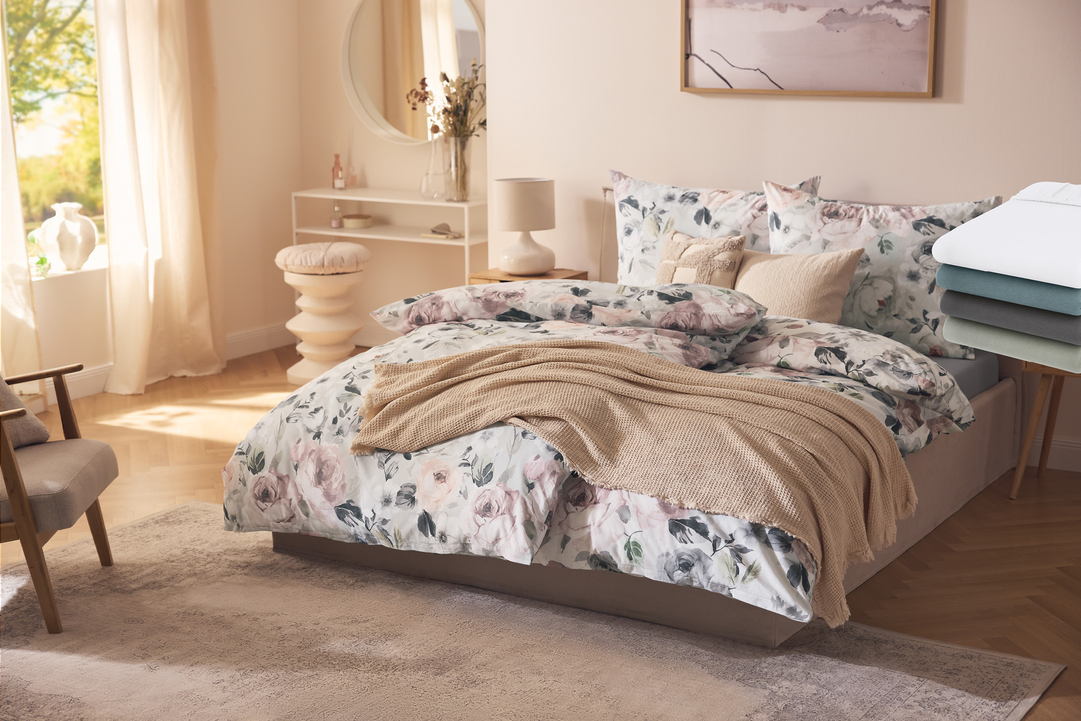 Blommiga sängkläder med en beige filt, omgiven av sovrumsinredning och en stapel enfärgade lakan.