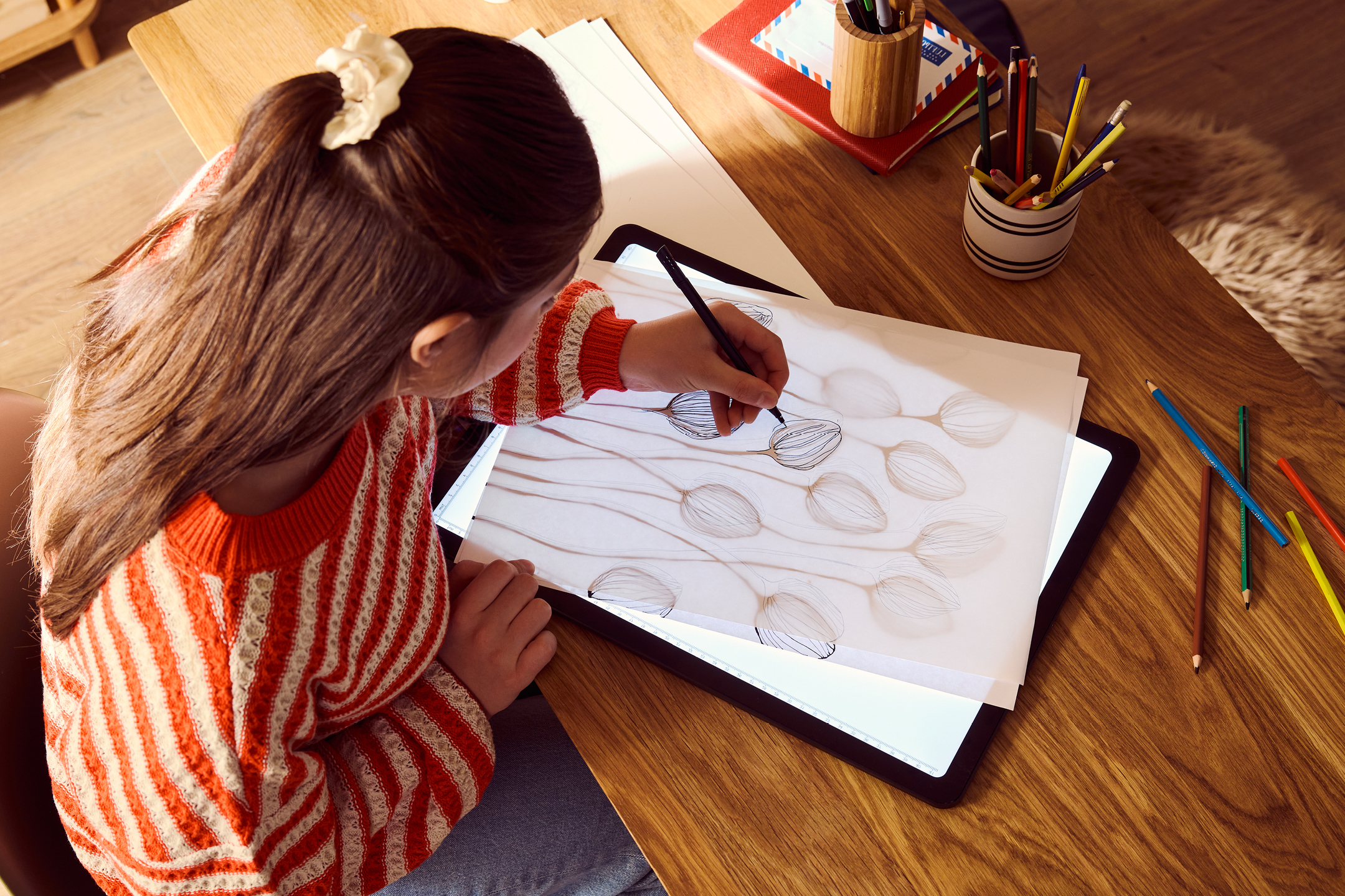 En person ritar på ett ritbord med en penna, med färgpennor och anteckningsböcker i närheten.