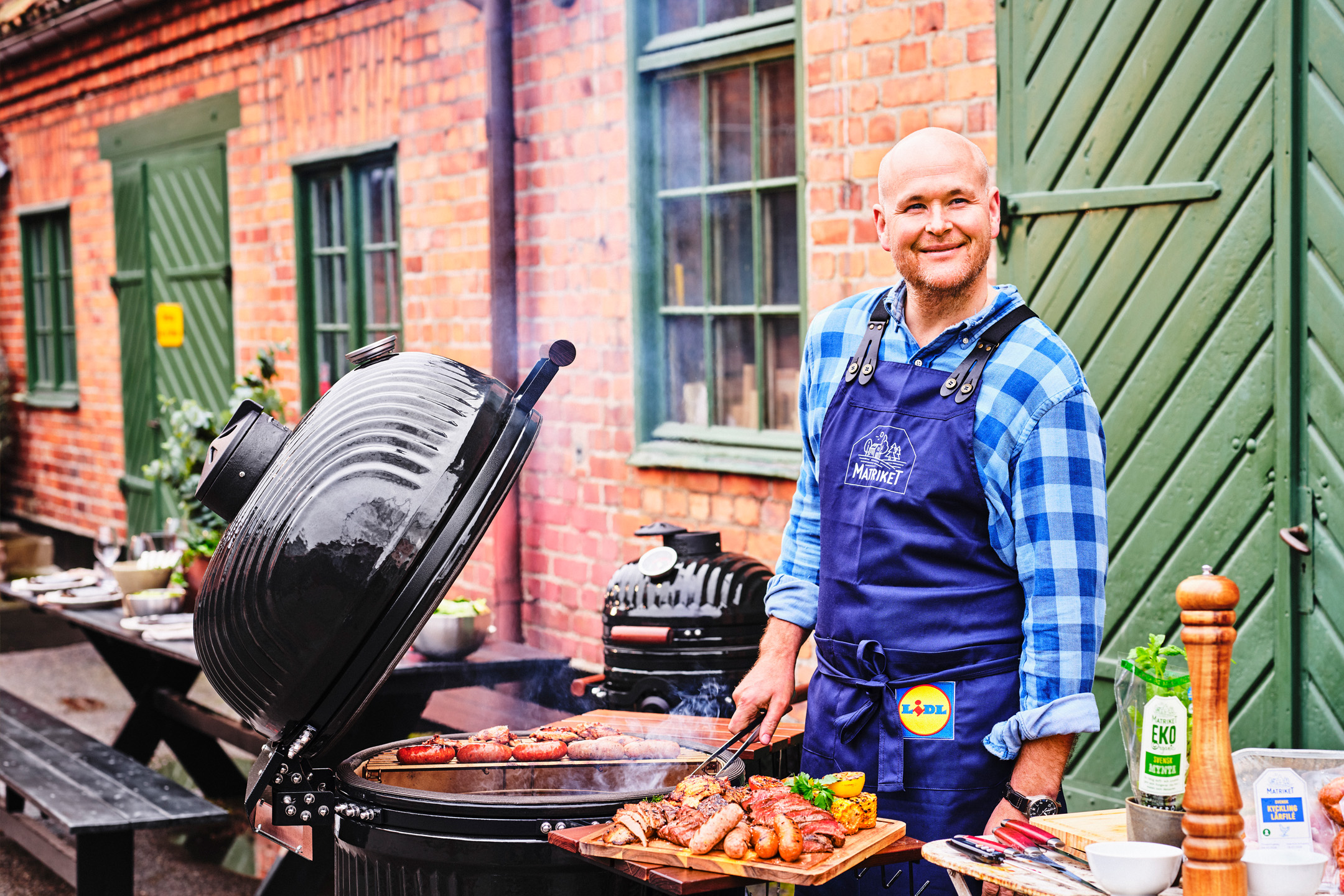 Man grillar korv och kött utomhus, med en grill och ett uppdukat bord i bakgrunden.