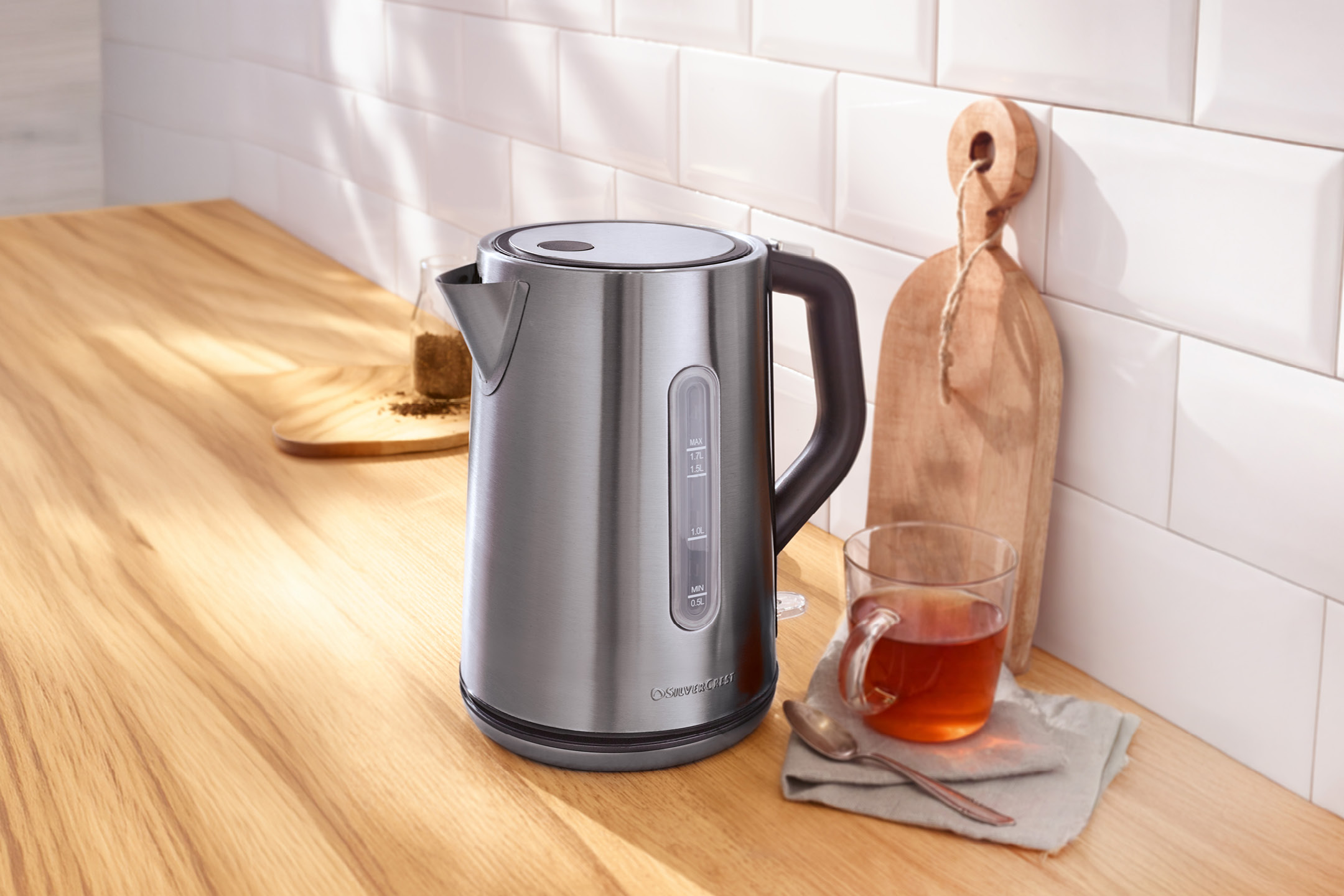 En silverfärgad vattenkokare på en träbänk med en kopp te och en skärbräda.