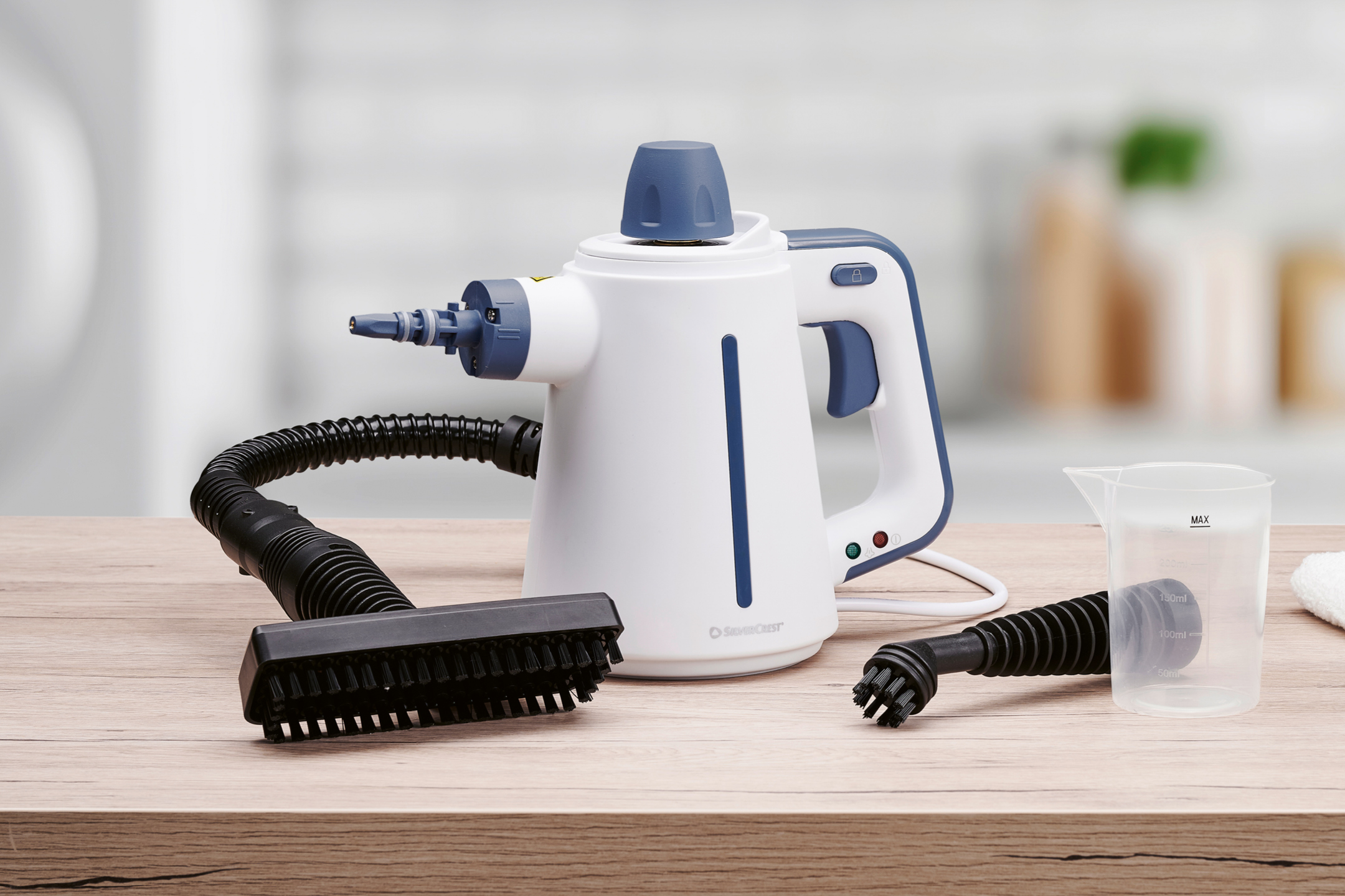 Handhållen ångtvätt med tillbehör som borstar och mätkopp på ett träbord.