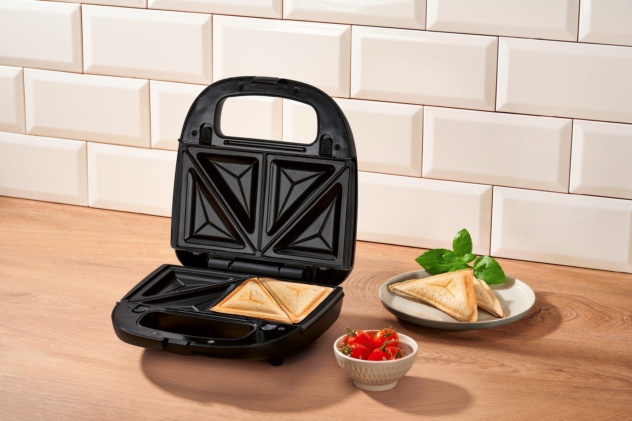 Öppen smörgåsgrill med en färdig toast, en tallrik med två toast och en skål med körsbärstomater.