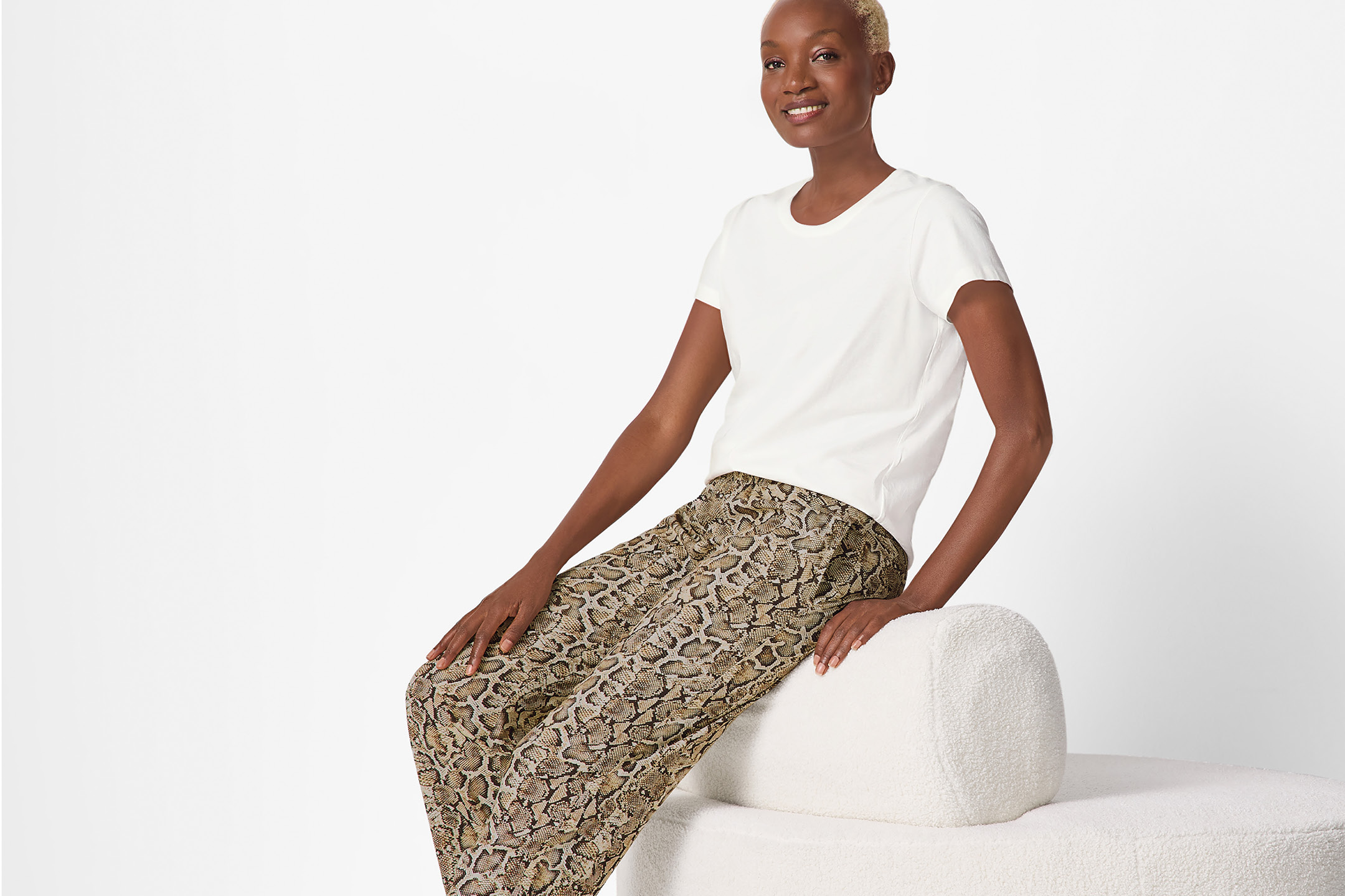 Kvinna i vit t-shirt och ormskinnsmönstrade byxor sitter på en vit bänk.