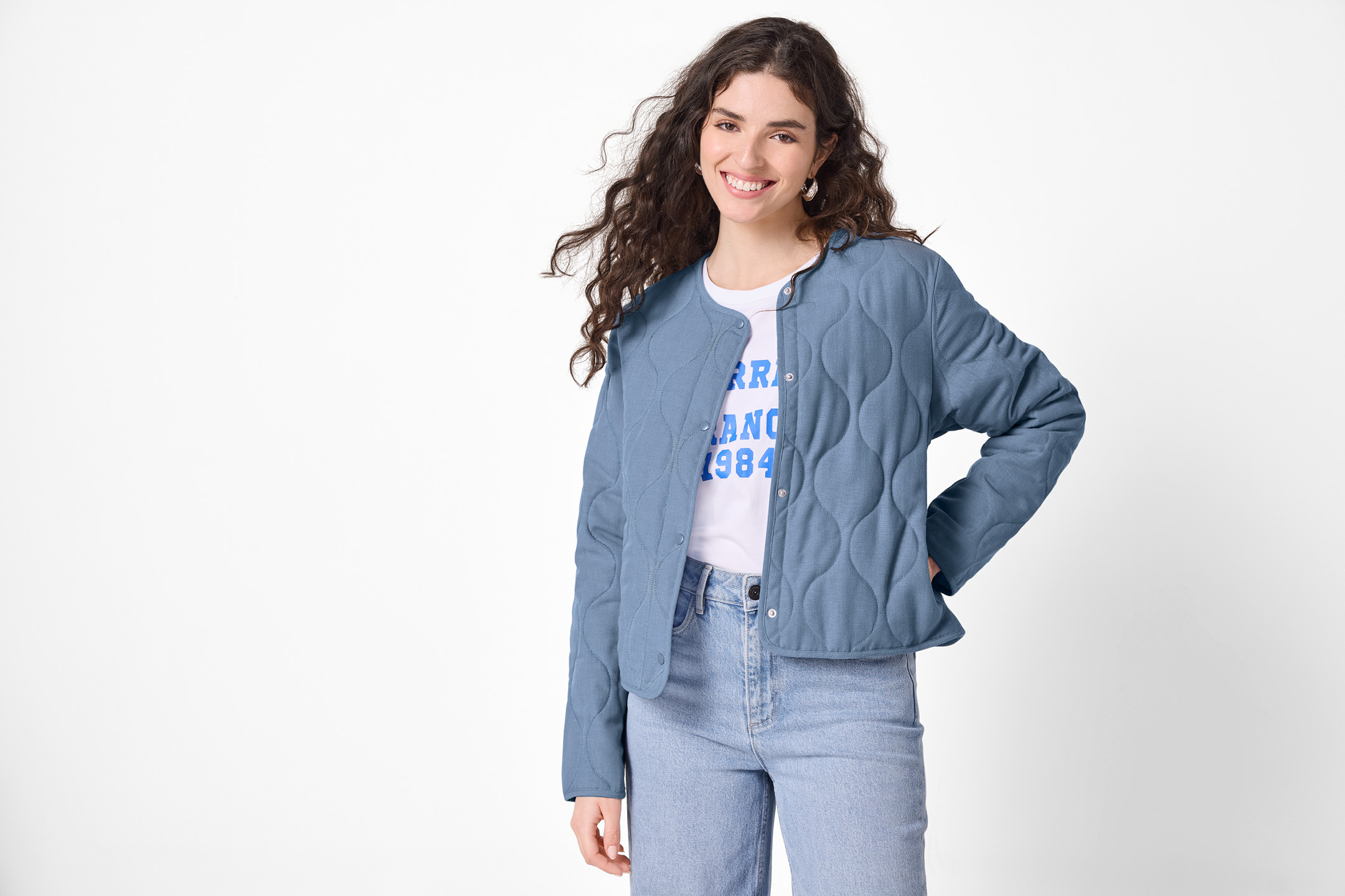 Kvinna i blå kviltad jacka, vit t-shirt med tryck och jeans.