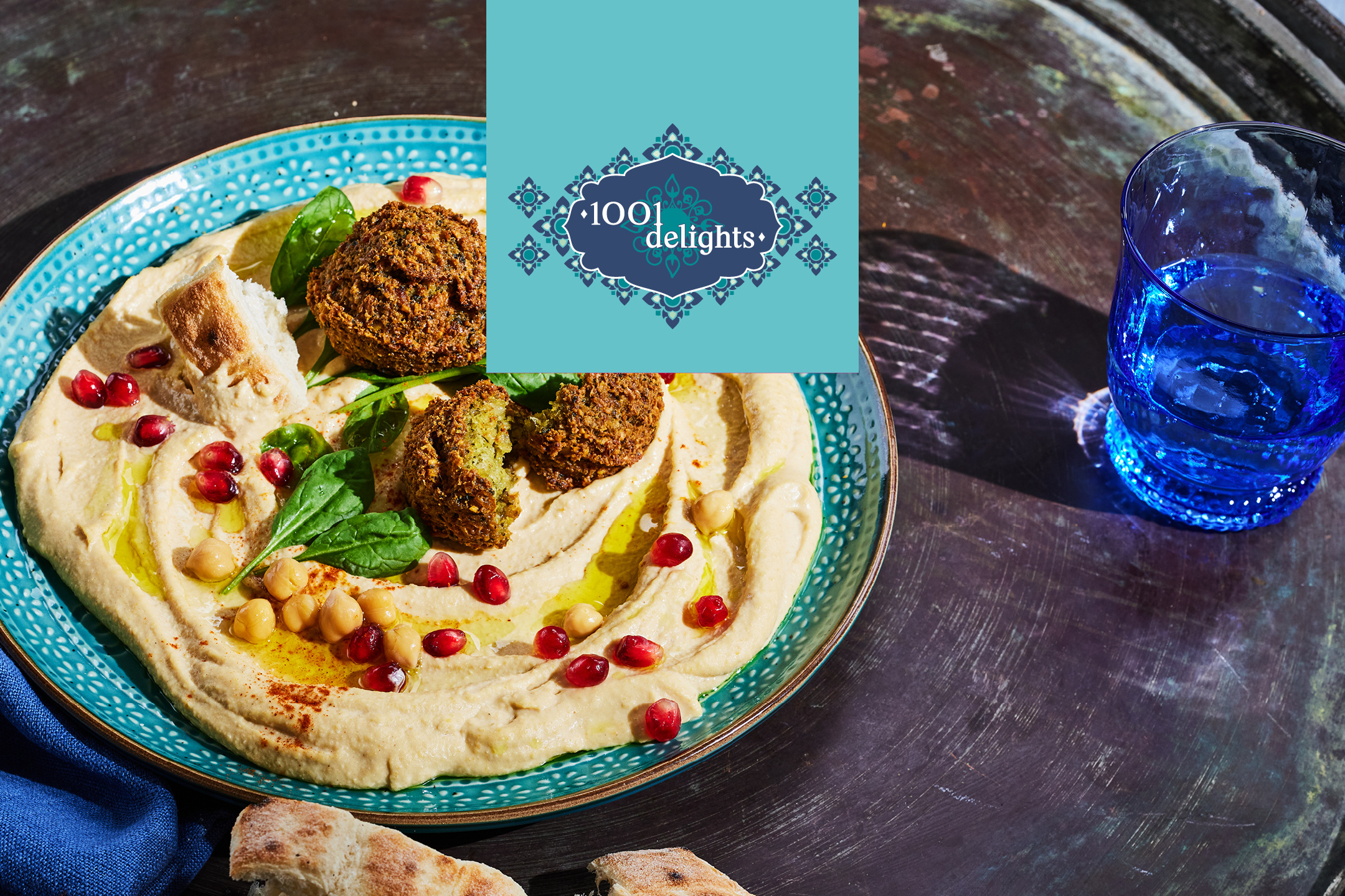 Hummus med falafel, granatäpple, kikärtor och bröd på en blå tallrik, med en blå vattenkopp.