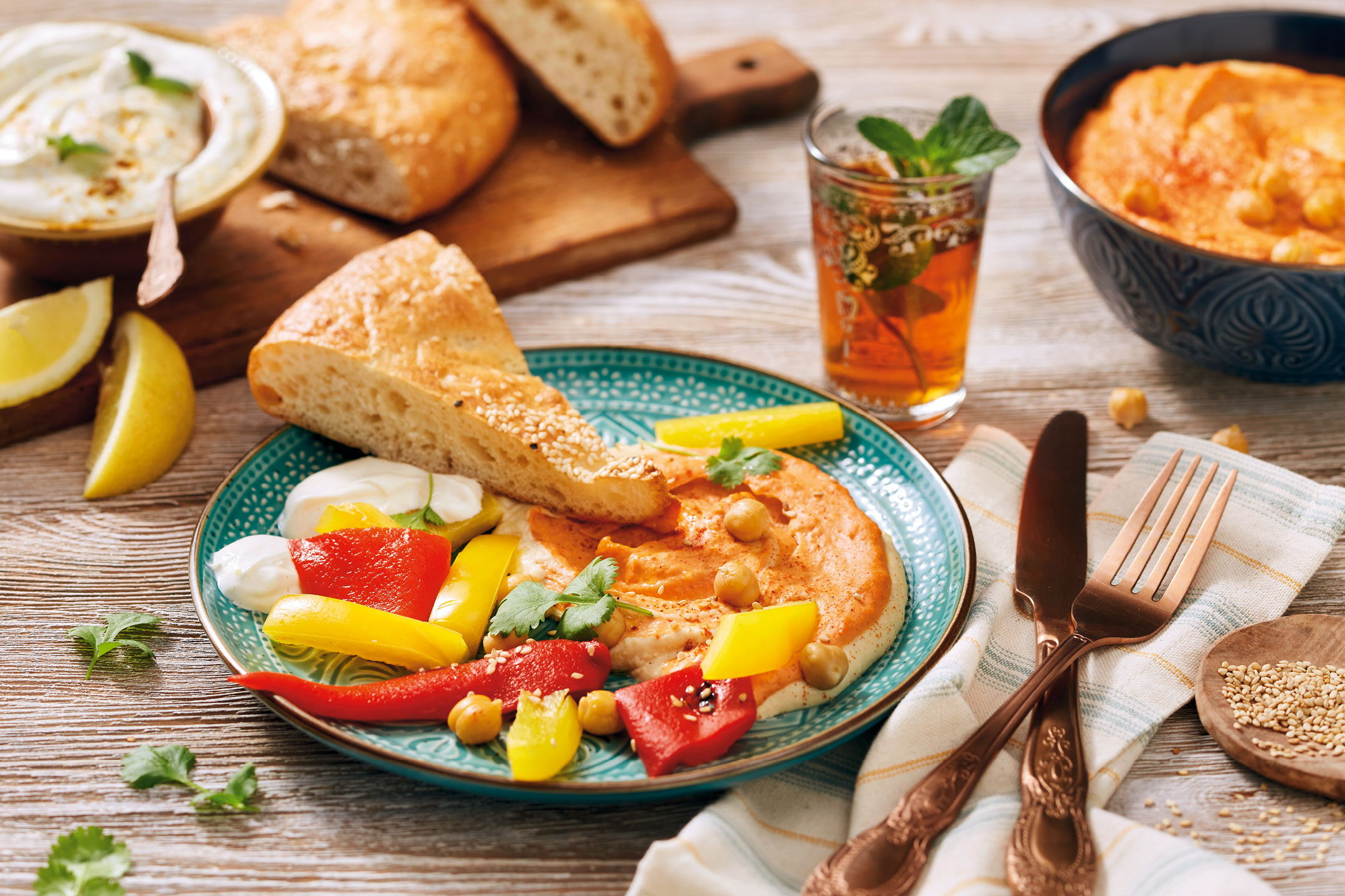 Hummus med paprika, bröd, yoghurt och myntate på ett träbord.
