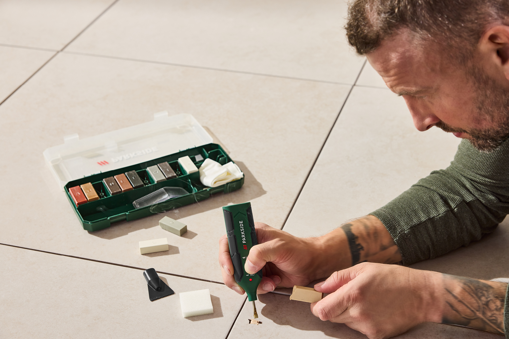 Man reparerar ett kakelgolv med ett reparationskit för kakel, inklusive en smältpenna och vaxkritor.