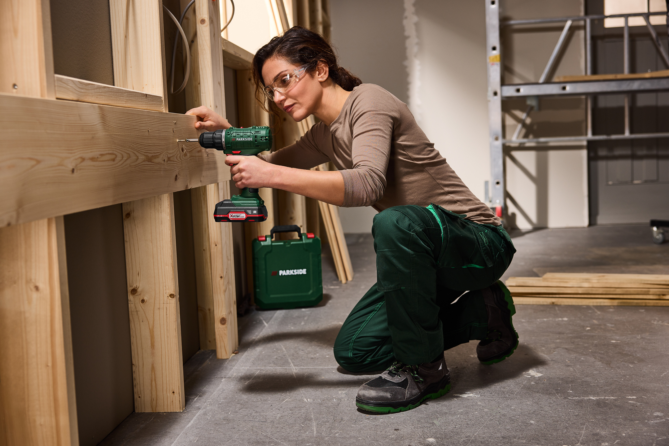 Kvinna i skyddsglasögon borrar i träregel med sladdlös borrmaskin, grön verktygslåda på golvet.
