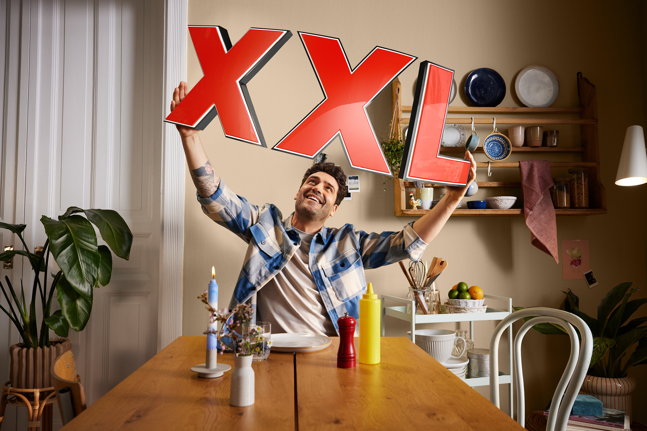 Man håller upp stora röda bokstäver som bildar 'XXL' i ett kök med hyllor och porslin.