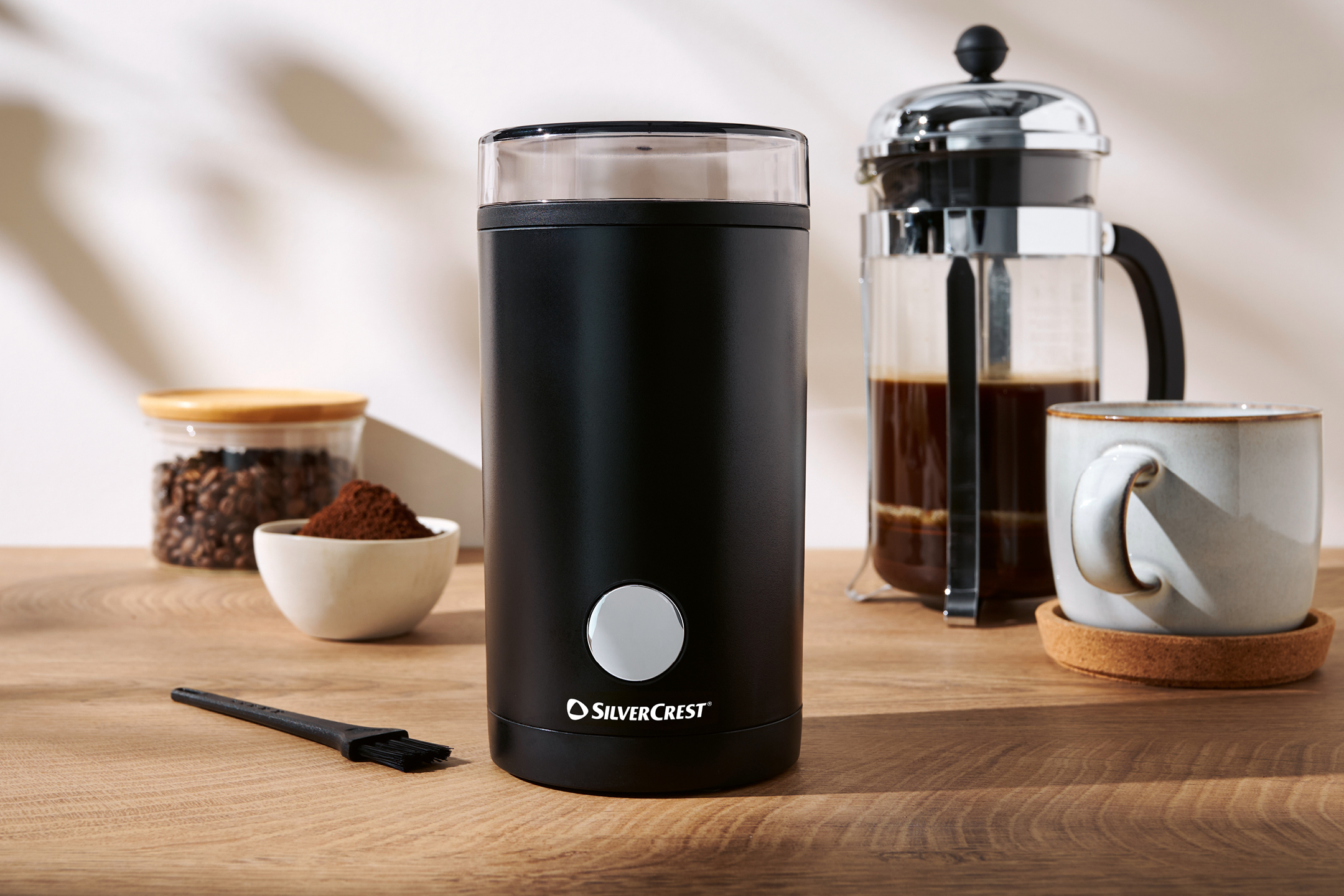 Svart kaffekvarn med kaffebönor, malt kaffe, en presskanna och en kaffekopp på ett träbord.