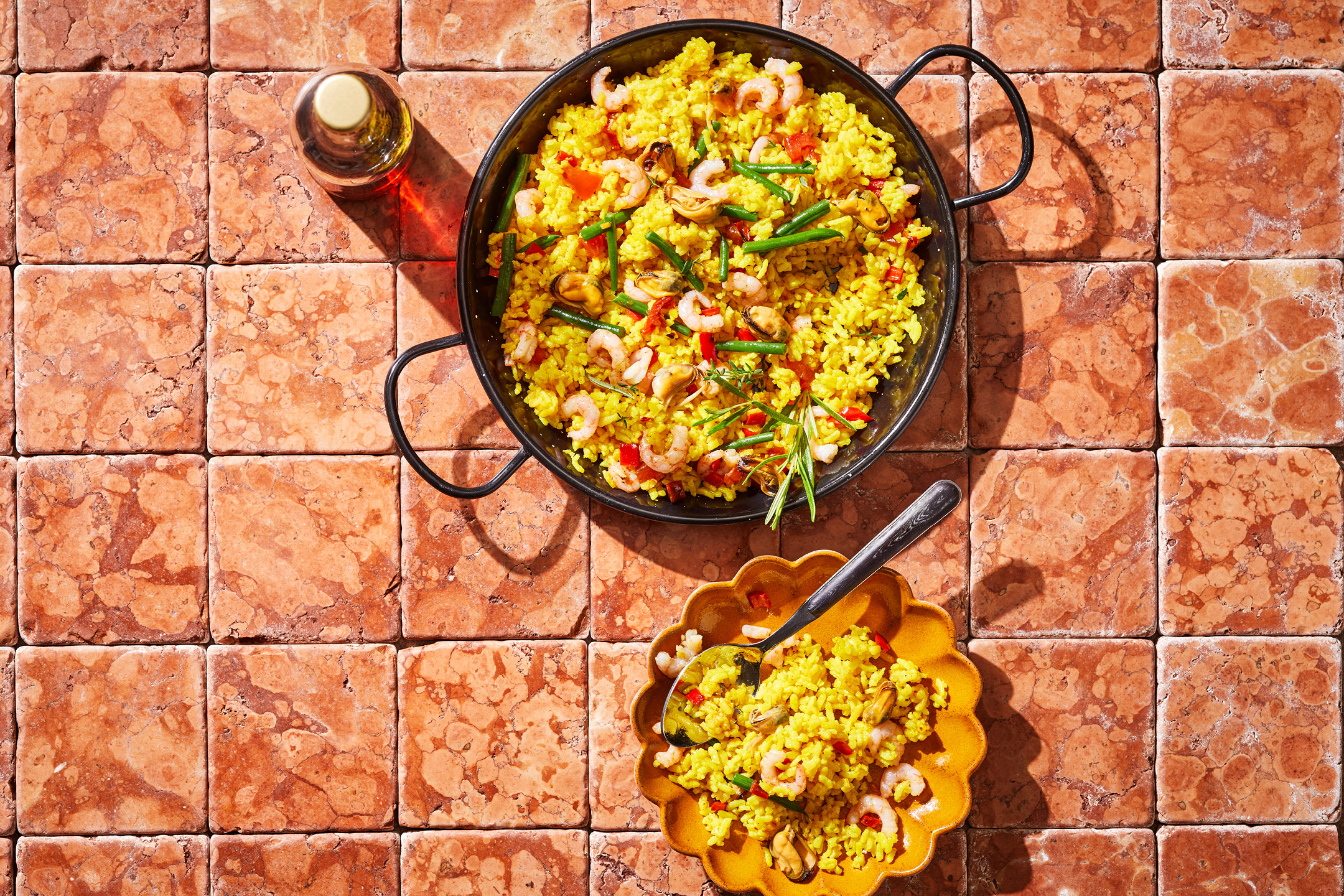 Paella med skaldjur, grönsaker och ris i en stor panna och en mindre tallrik på ett kaklat bord.