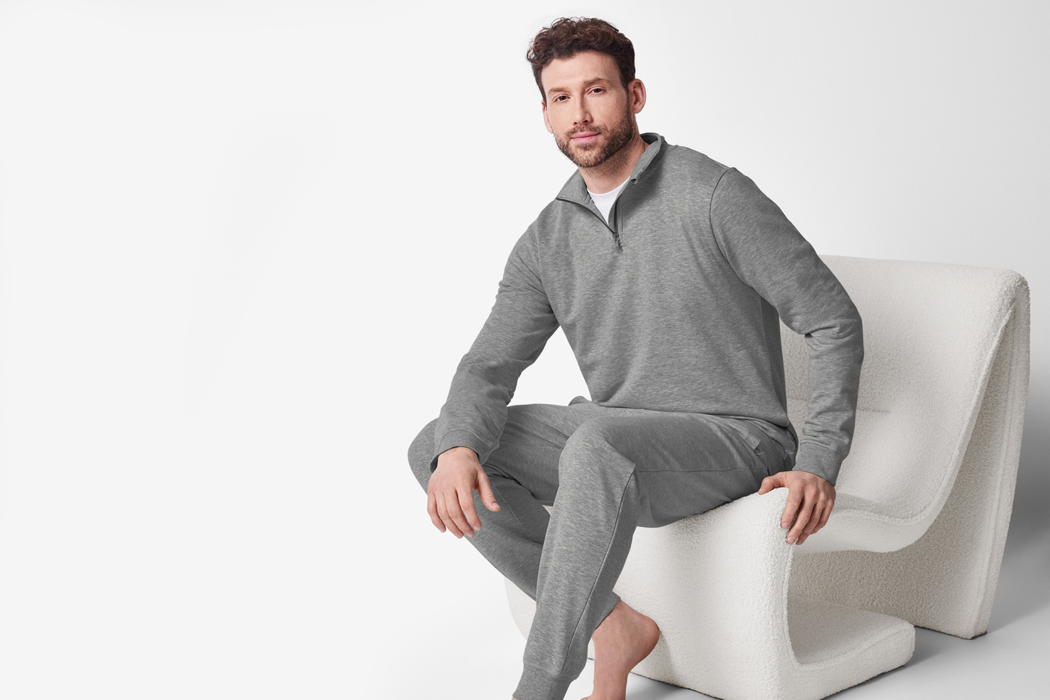 Man i grå mysdress med tröja med dragkedja och matchande byxor, sittandes i en vit fåtölj.