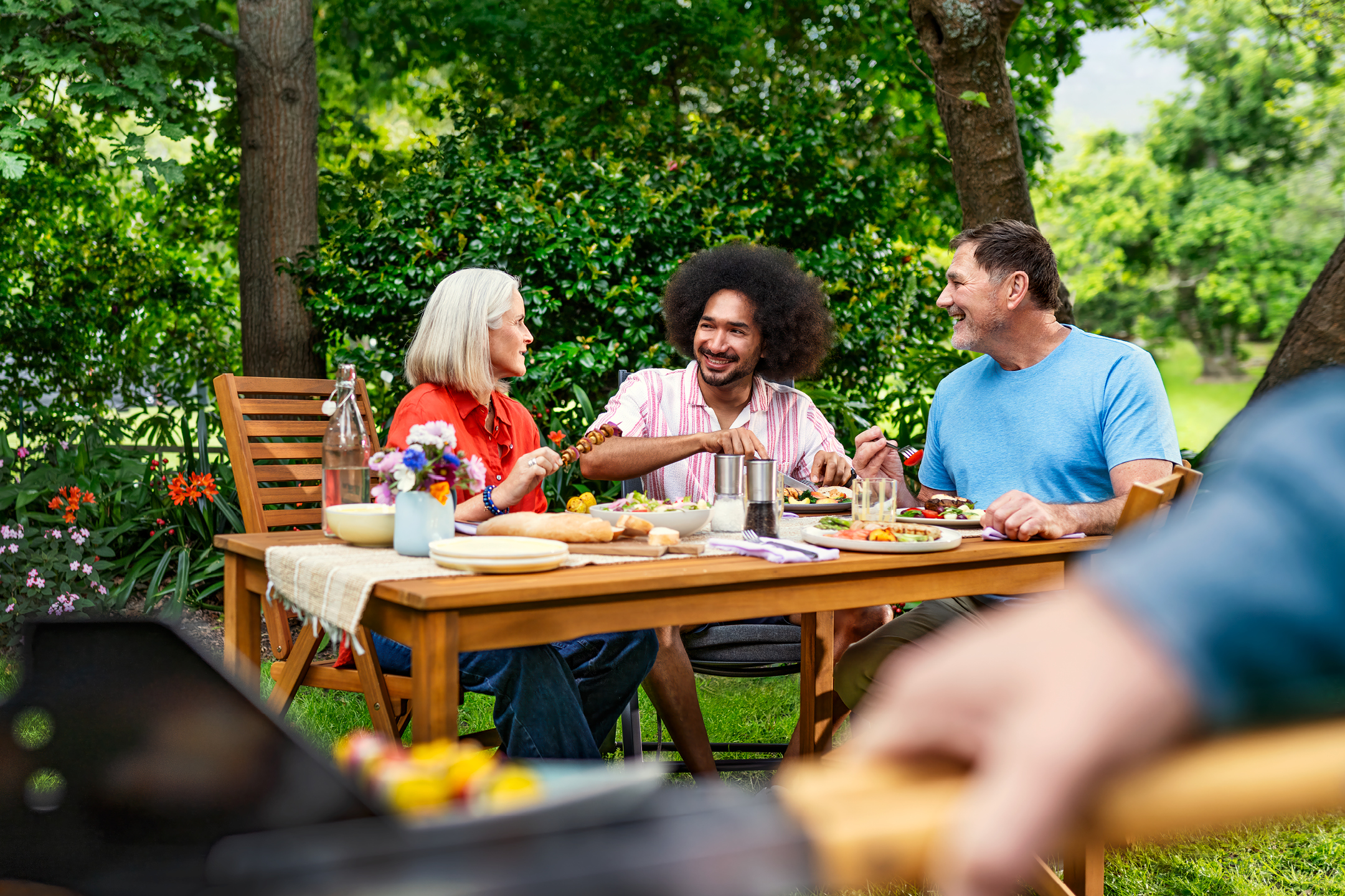 Vänner grillar och äter utomhus vid ett träbord med mat och dryck.