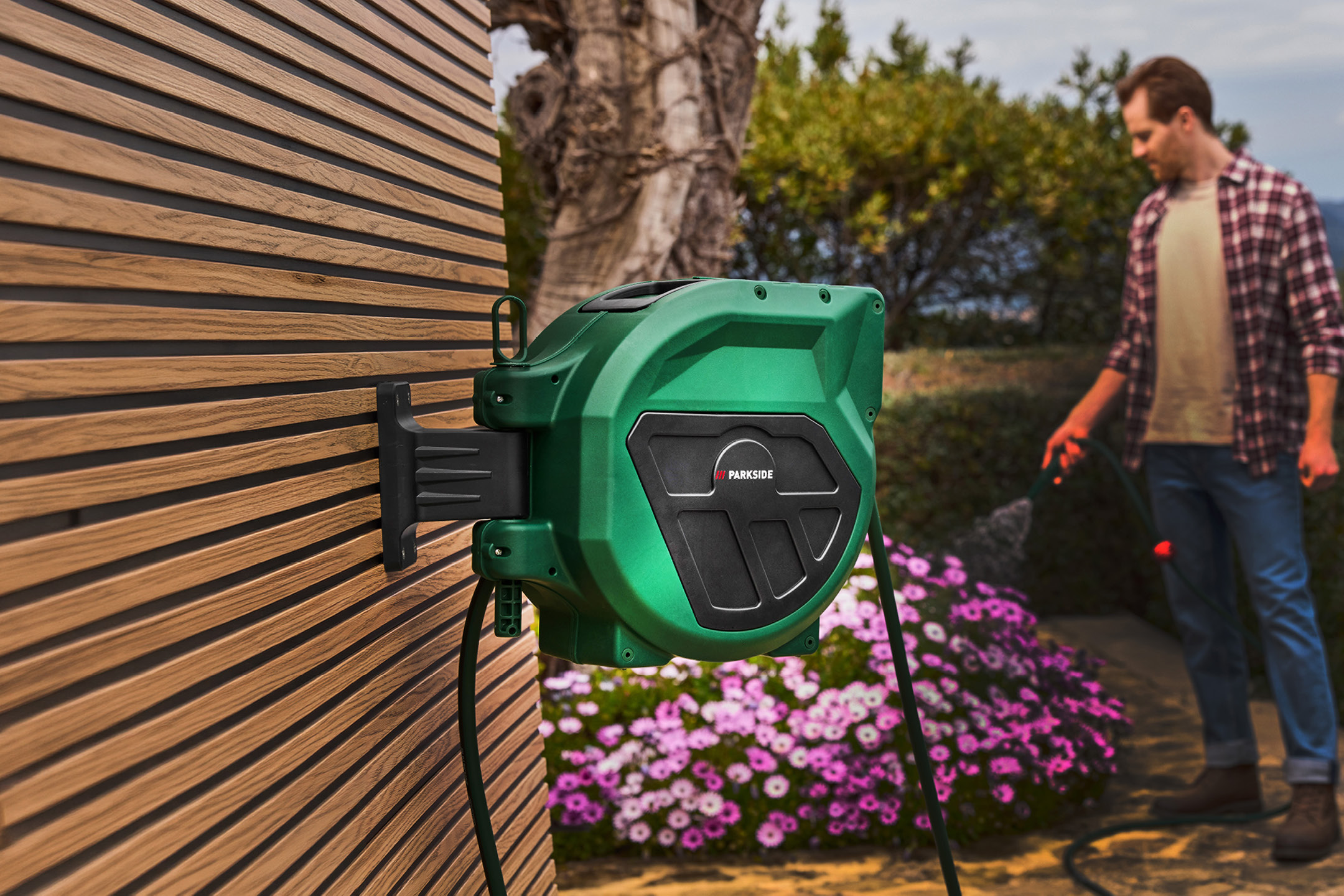 Grön slangvinda monterad på en träpanelvägg, med en man som vattnar blommor i bakgrunden.