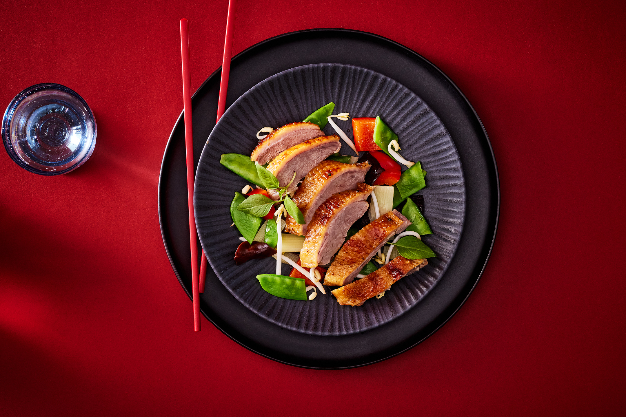 Anka med grönsaker och böngroddar på en svart tallrik med röda ätpinnar och ett glas.