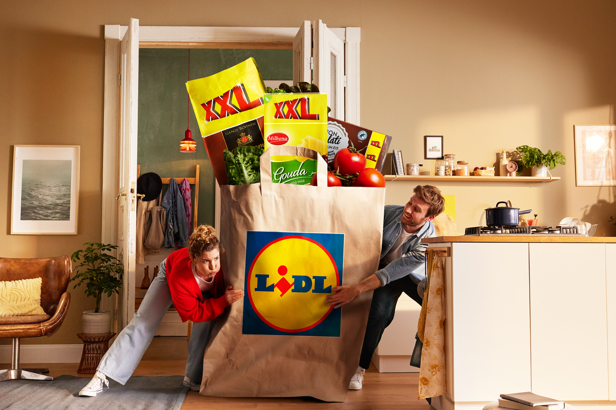 Två personer bär en stor matkasse fylld med XXL-produkter, inklusive kaffe, ost, sallad och tomater.