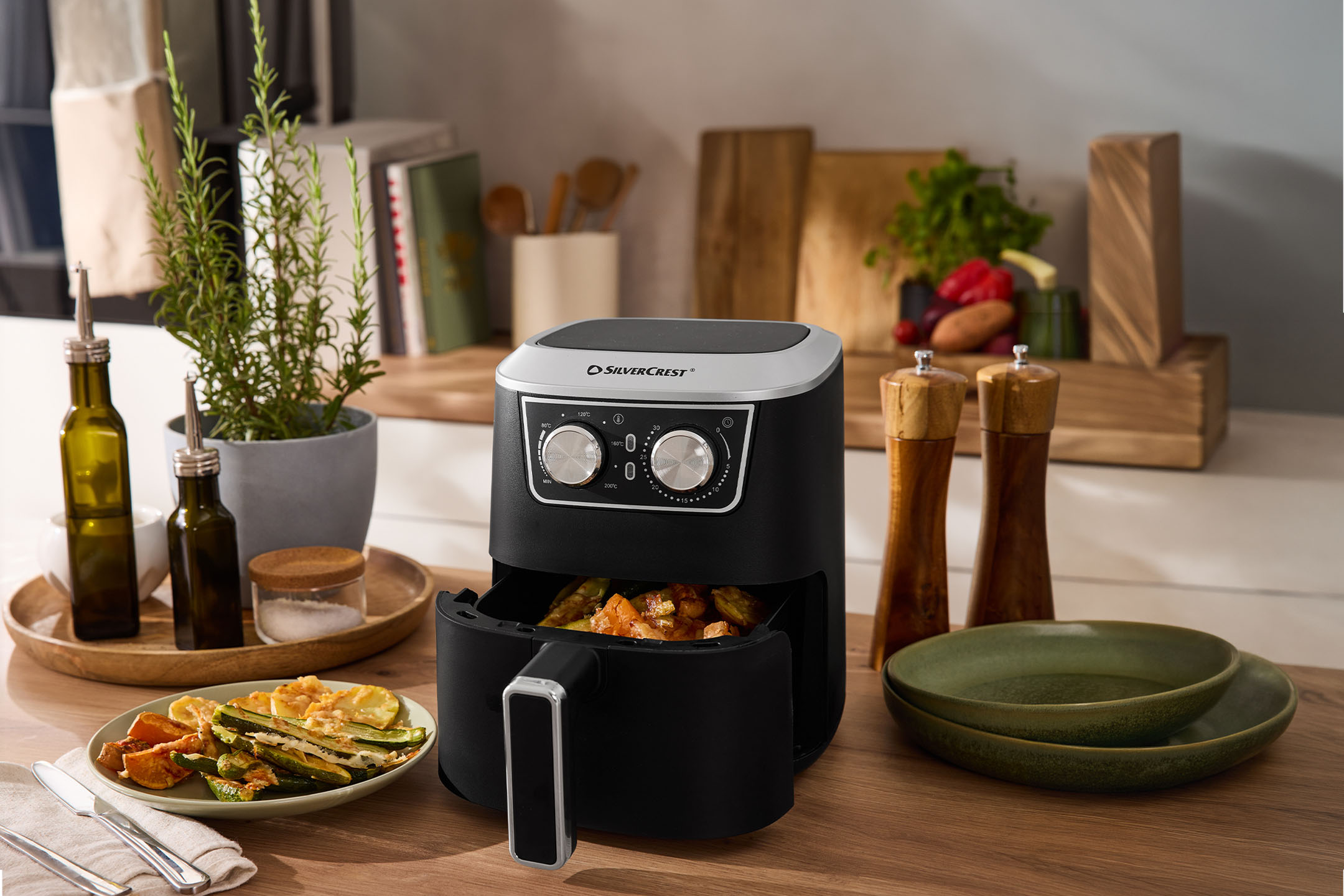 Svart airfryer med grönsaker, kryddor och tallrikar på en träbänkskiva.