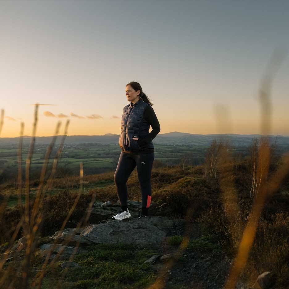 Kvinna i vadderad väst och leggings står på en bergstopp vid solnedgången.