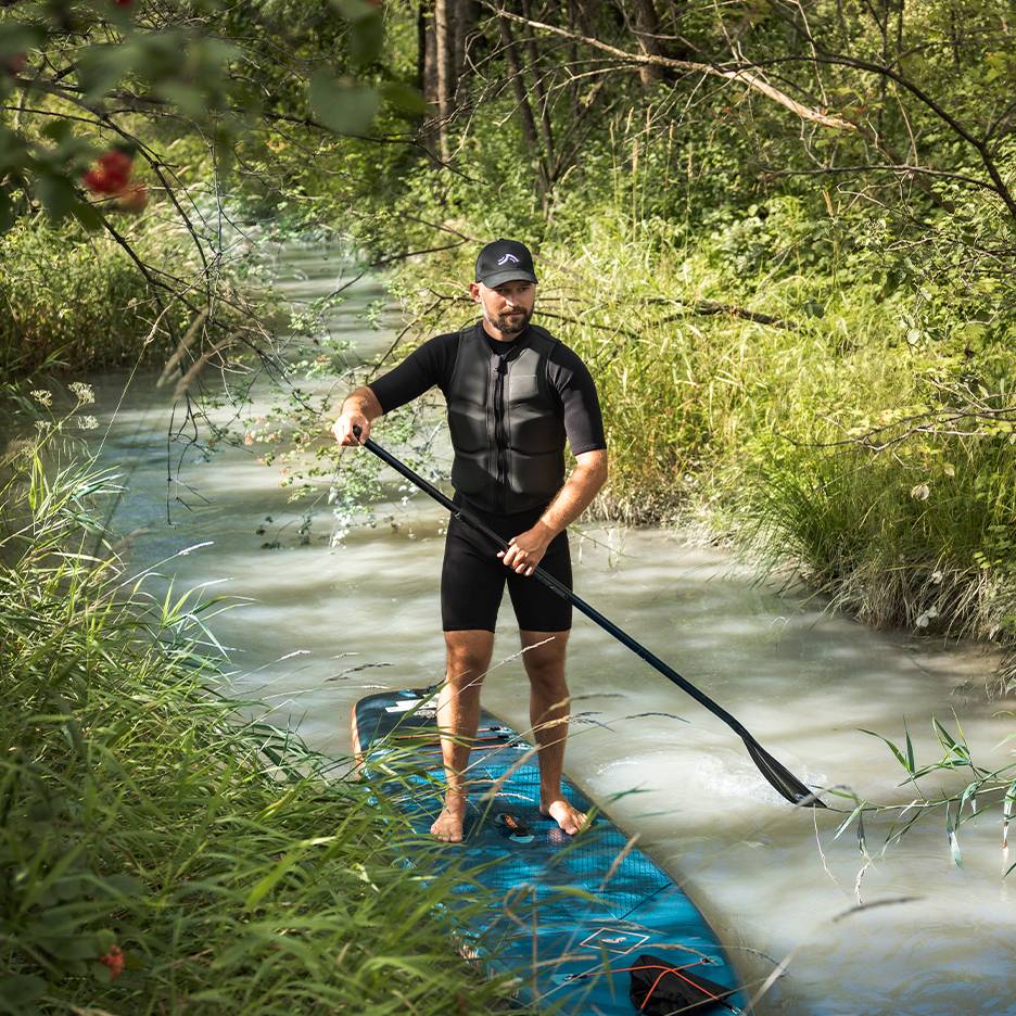 Man i våtdräkt och flytväst står på en paddleboard i en bäck.