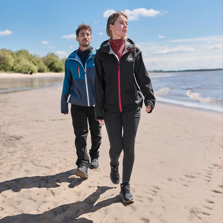 Par i vandringsjackor och byxor går på en sandstrand vid havet.