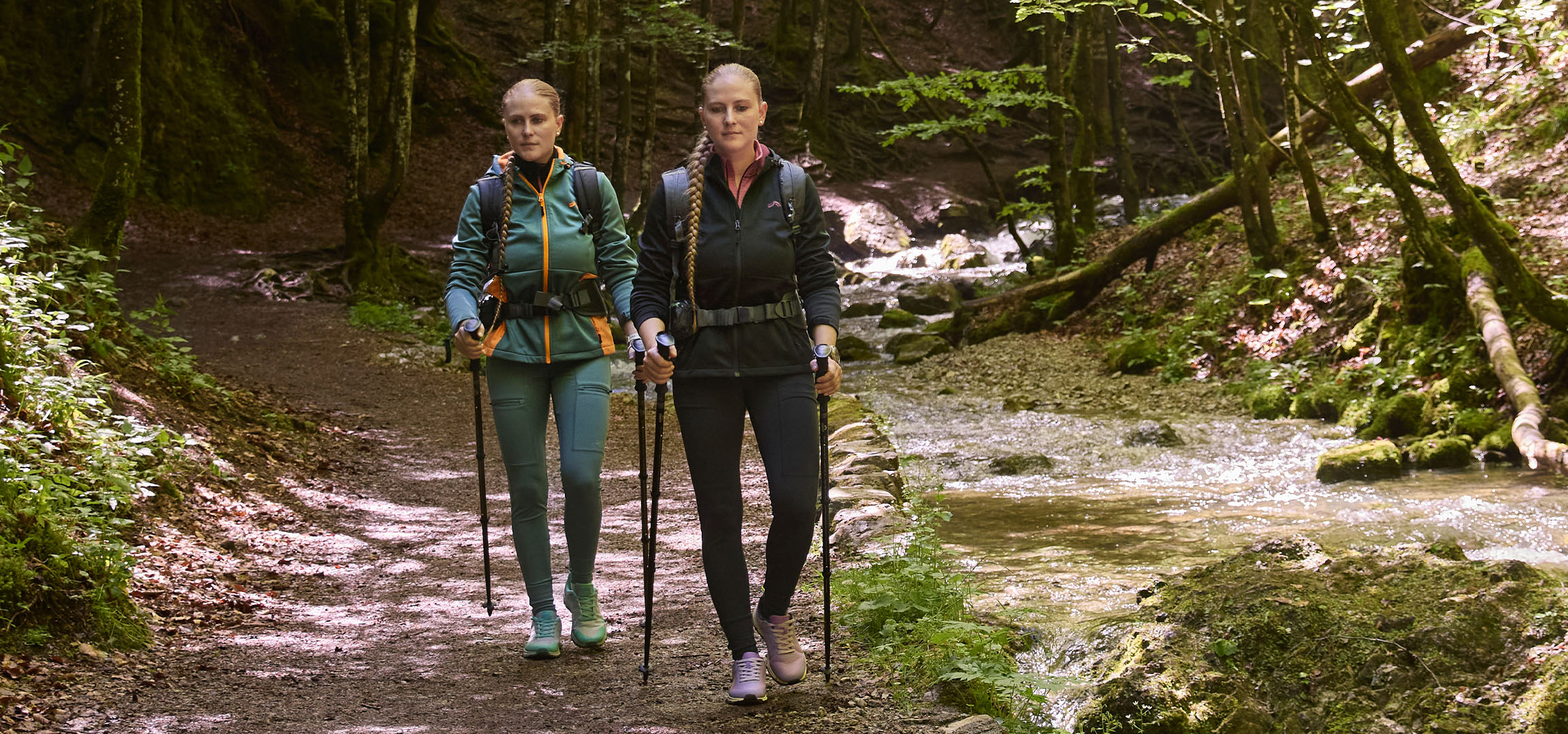 Två kvinnor vandrar i en skog längs en bäck med vandringsstavar.