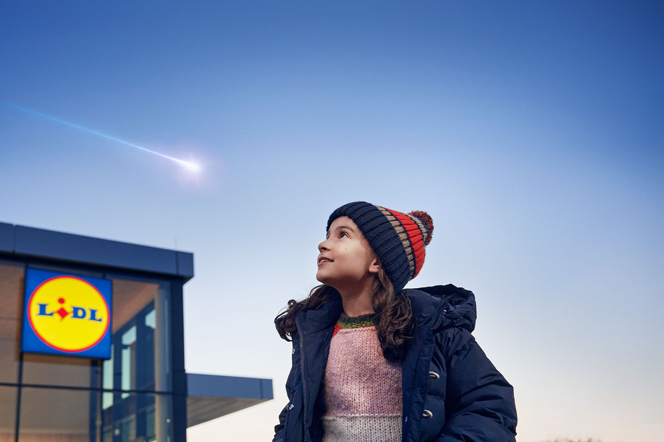 Flicka i mössa tittar på en meteor på himlen, med en Lidl-butik i bakgrunden.