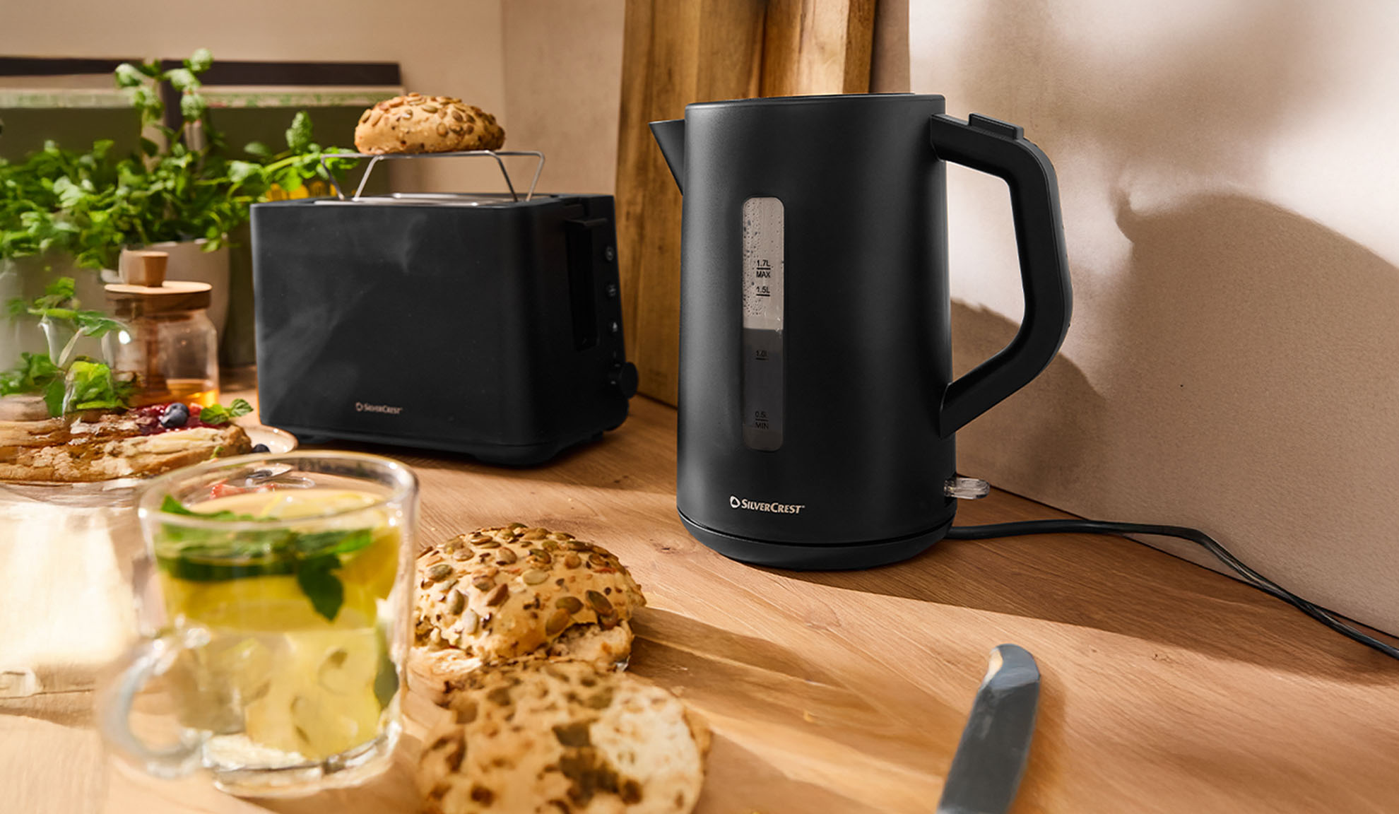 Svart SilverCrest vattenkokare och brödrost på ett träbord med frukost.