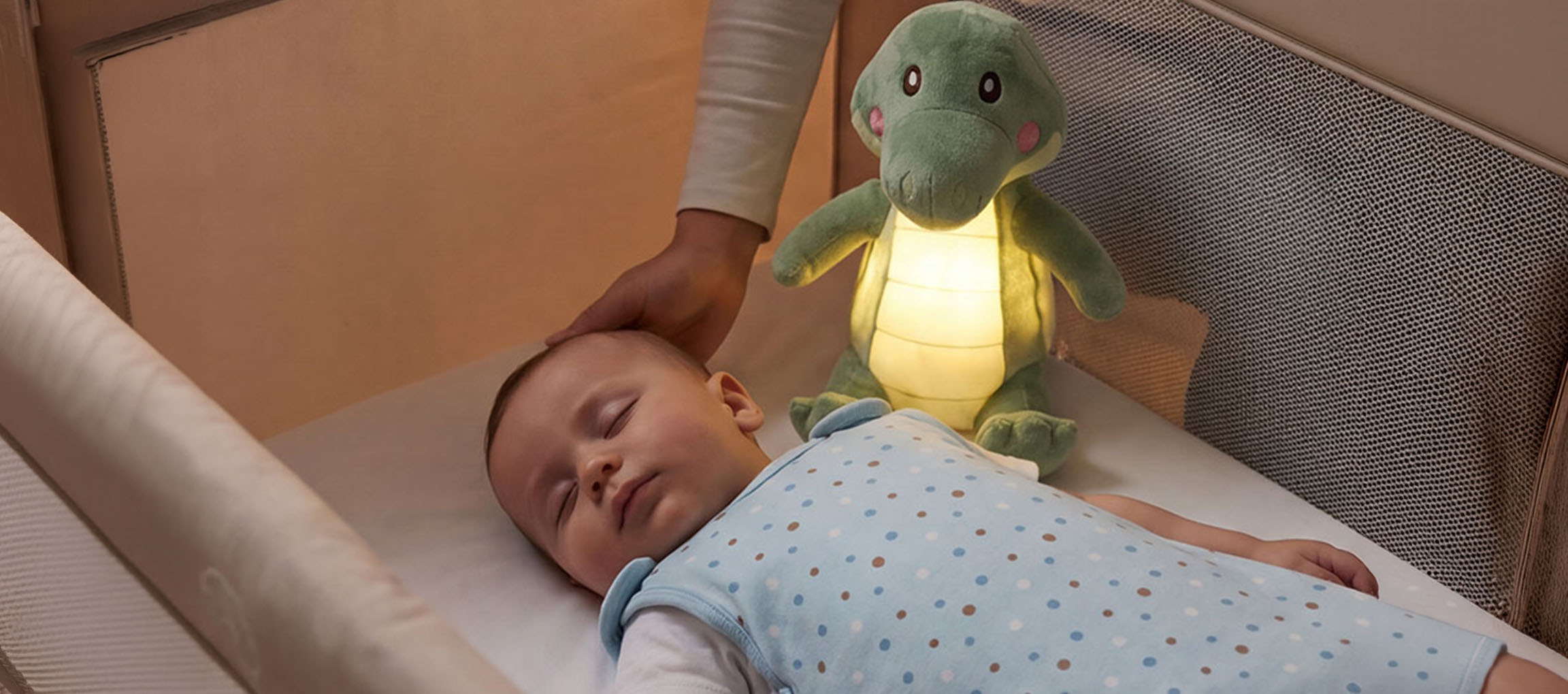 Baby sover i spjälsäng med gosedjursnattlampa, vuxen klappar barnets huvud.