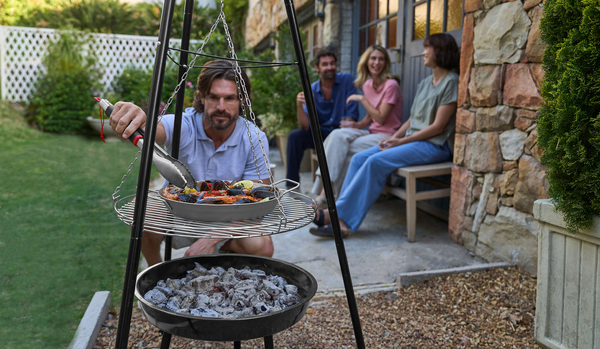 Man grillar skaldjur på stativ, vänner kopplar av i bakgrunden.