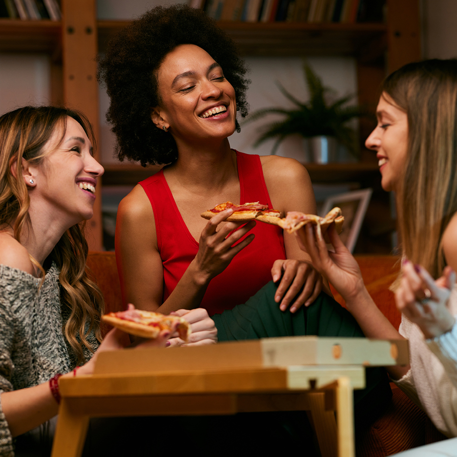 Tre glada kvinnor äter pizza tillsammans.