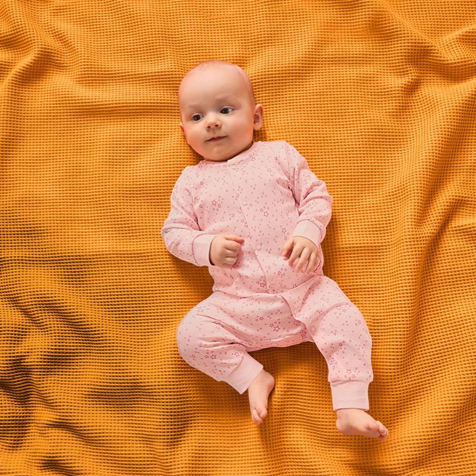 En bebis i en rosa mönstrad pyjamas ligger på en gul filt.