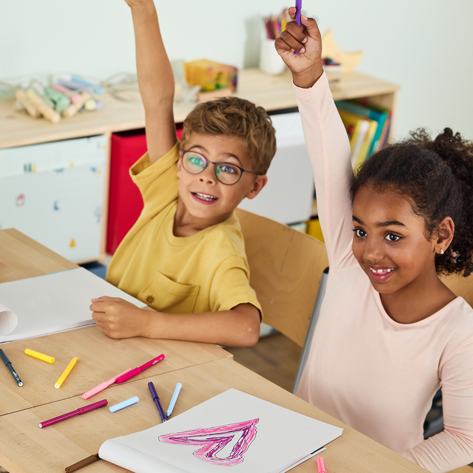 Glada barn ritar med färgpennor vid ett bord och räcker upp händerna.