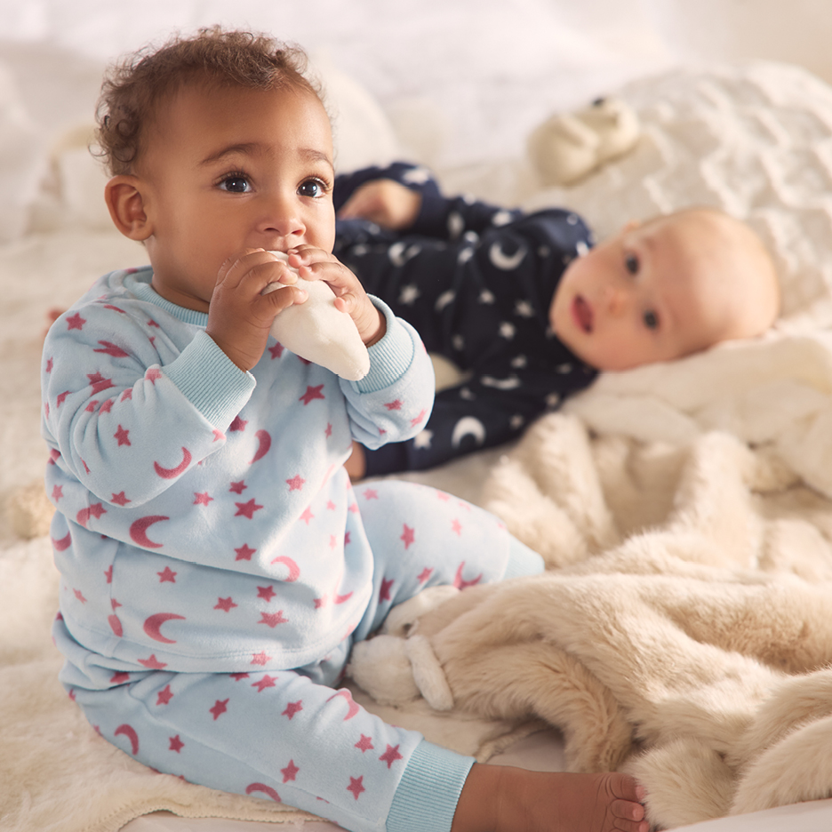 Två bebisar i pyjamas med stjärnor och månar, en tuggar på en vit leksak.