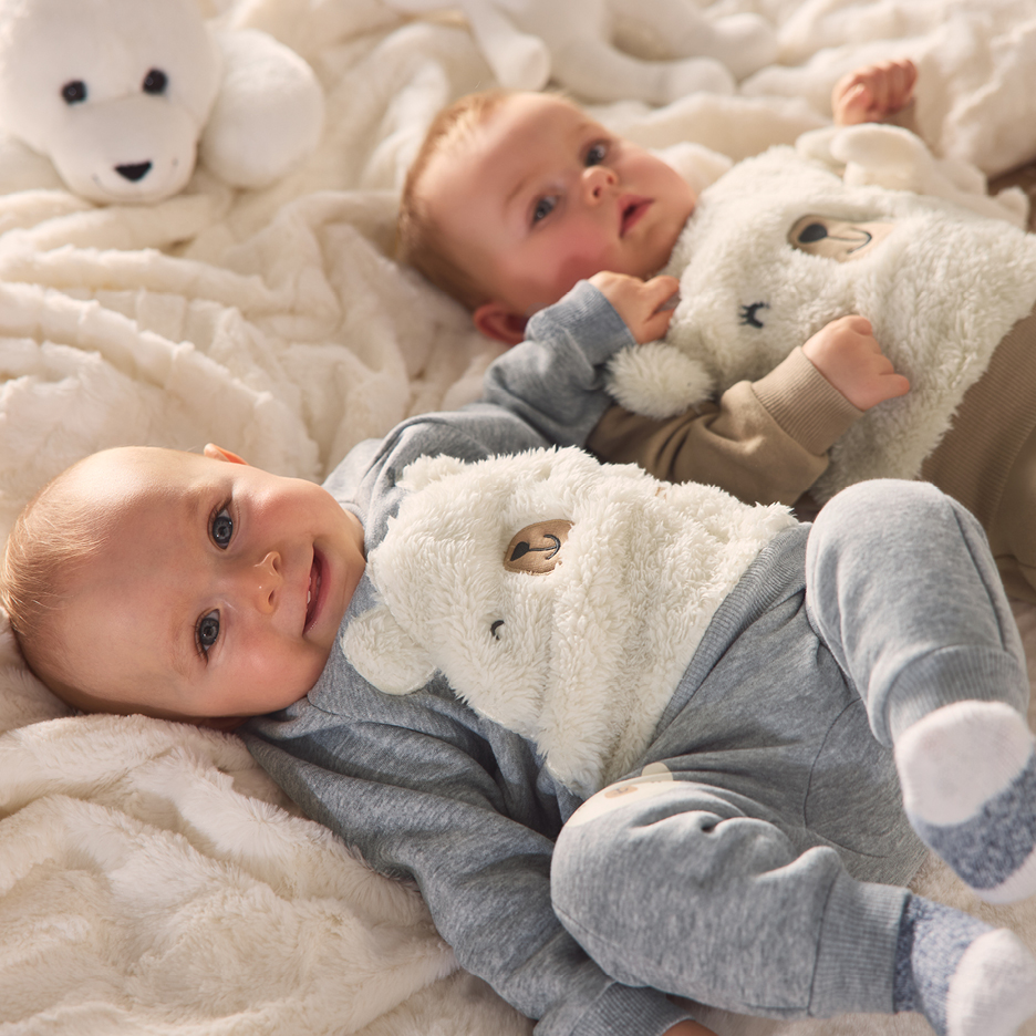 Två bebisar i mysiga björndräkter ligger på en mjuk, vit filt med gosedjur.