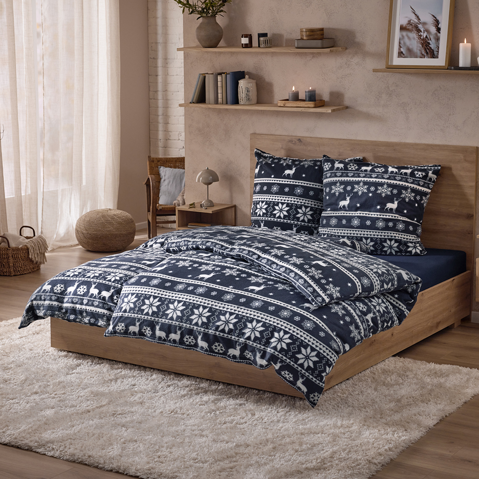 Säng med mörkblå sängkläder med snöflingor och renar, bredvid en puff och ett sängbord.