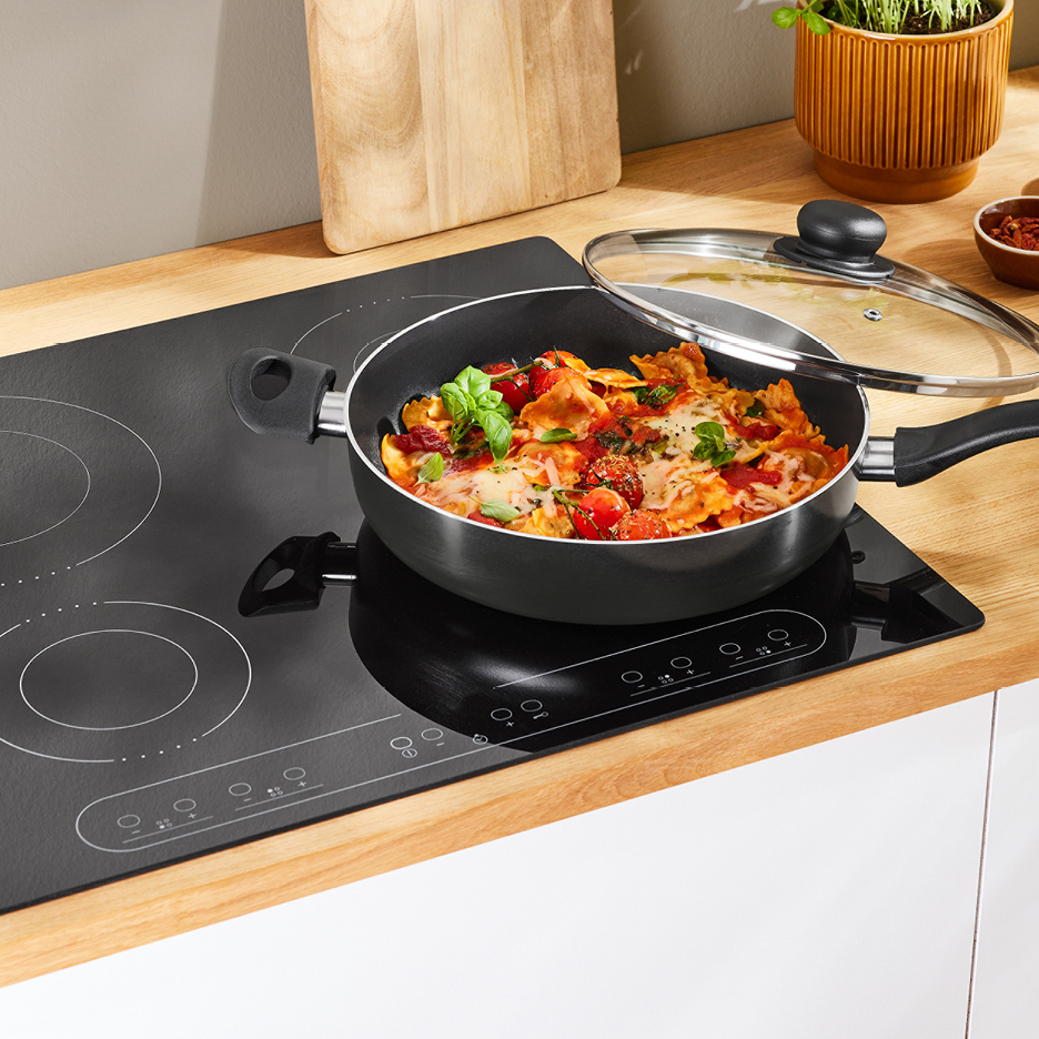 Svart induktionshäll med en stekpanna fylld med pasta och tomater.