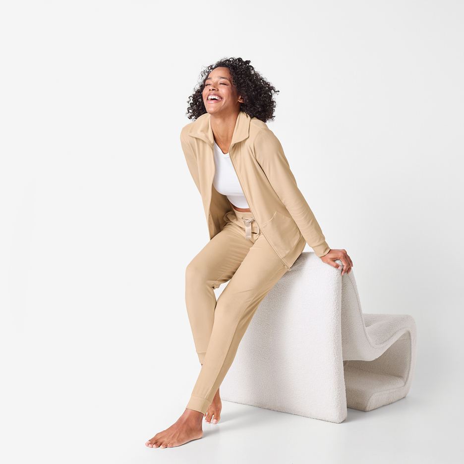 Kvinna i beige träningsset och vit topp, skrattar och sitter på en vit pall.