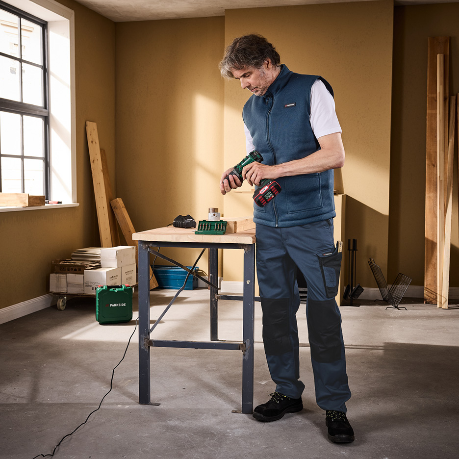 Man i arbetskläder med borrmaskin vid arbetsbänk i verkstad.