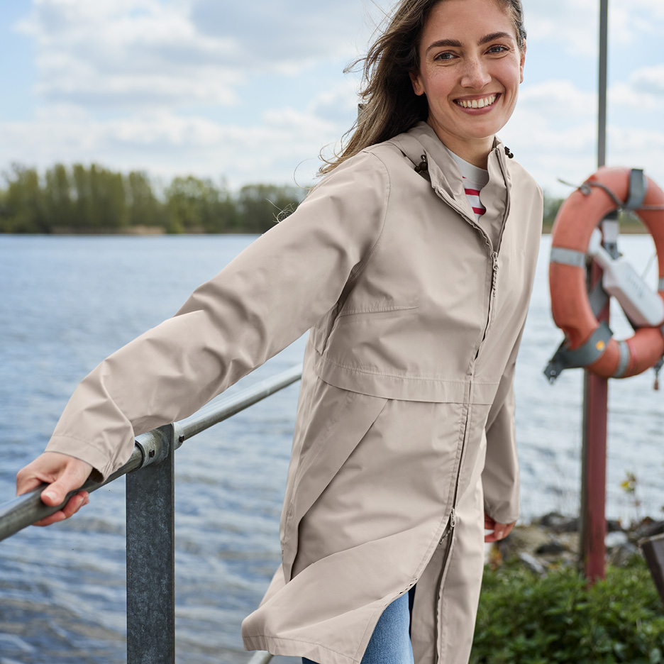 Kvinna i beige regnjacka och jeans vid en sjö, håller i ett räcke och ler.