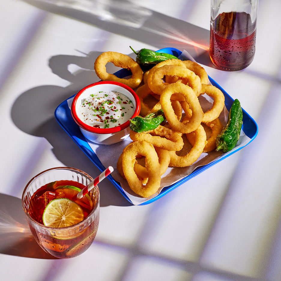 Friterade lökringar med dipp, padronpeppar och drinkar på ett vitt bord.