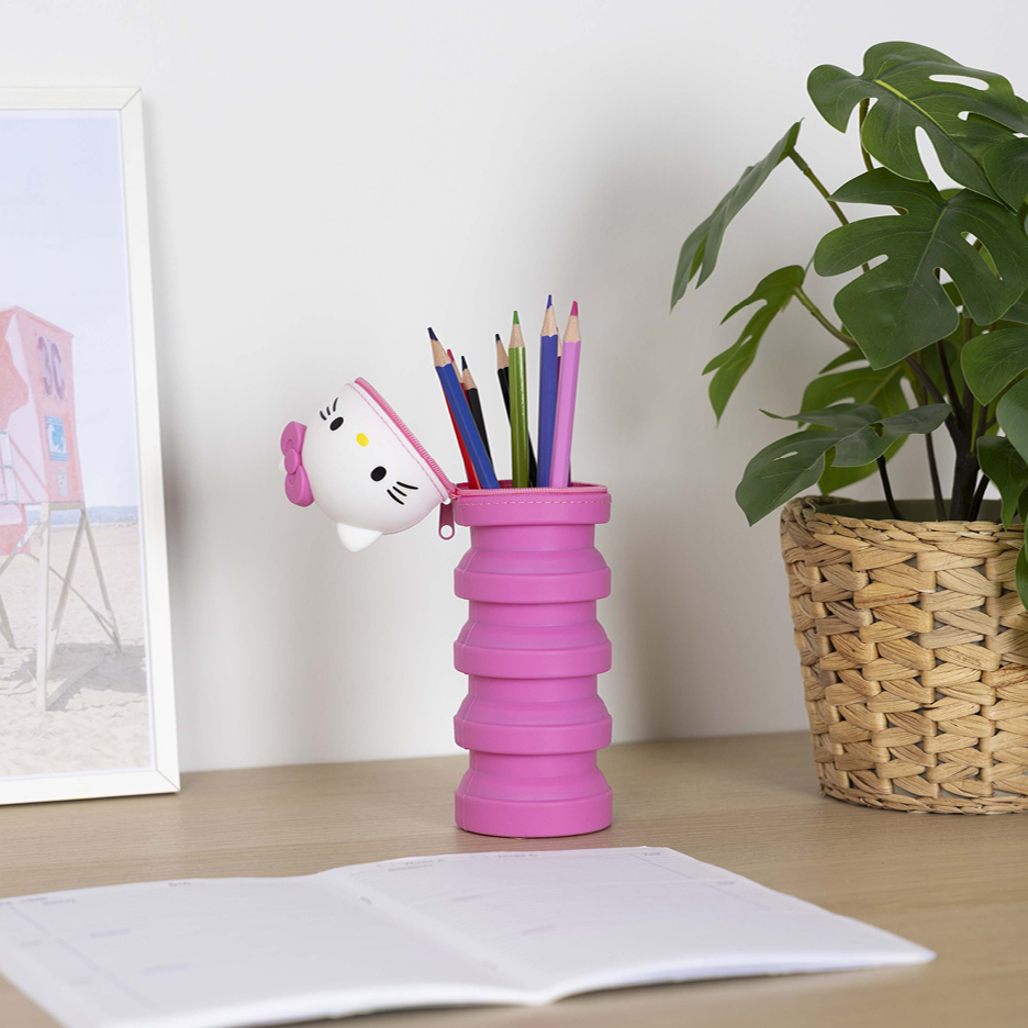 Rosa pennfodral med Hello Kitty-design, fyllt med färgpennor, på ett skrivbord med en växt och en anteckningsbok.