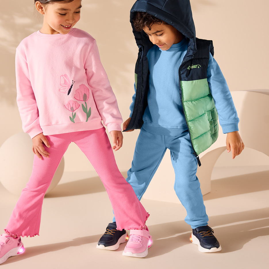 Barn i rosa tröja med fjäril och blommor, rosa byxor och blå tröja med blå byxor och grön väst.