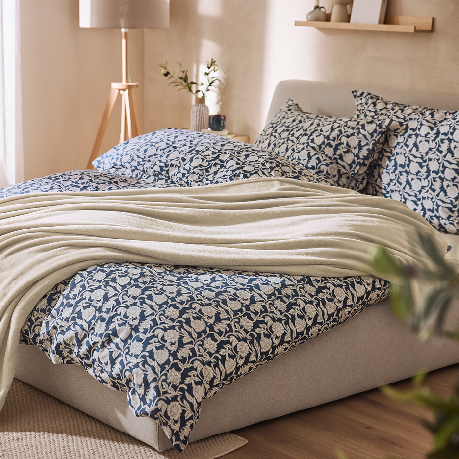 Säng med blått och vitt blommigt påslakan, matchande kuddar och en beige filt.